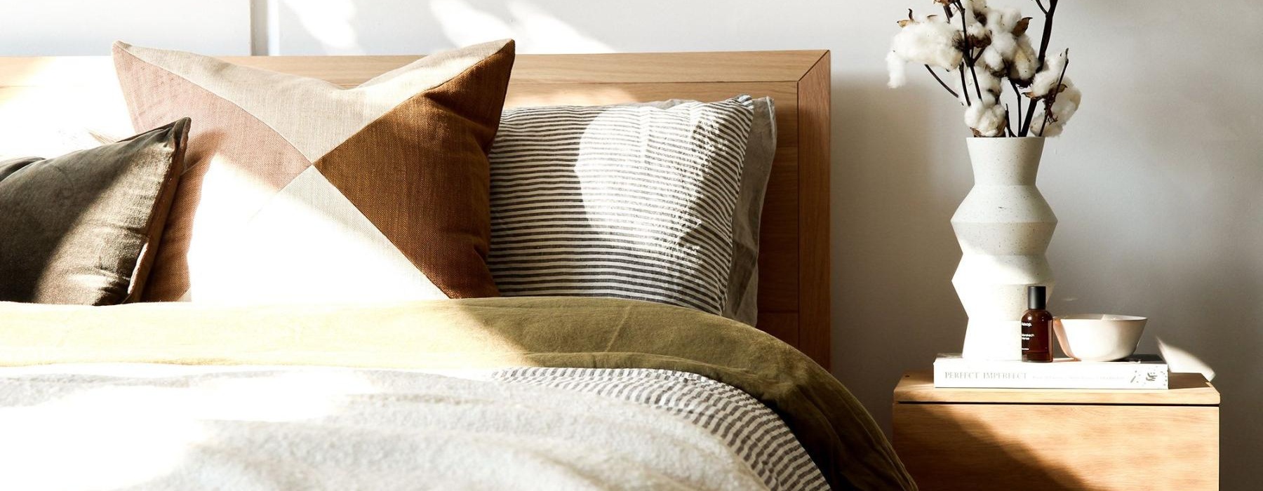 swath of sunlight shines across a well made bed and bedside table with a vase of cotton plants